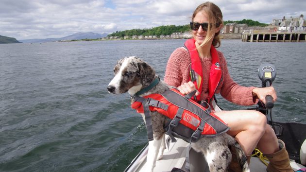 A woman wearing a red lifejacket operating an outboard engine, while a dog in a lifejacket leans over the side of a dinghy