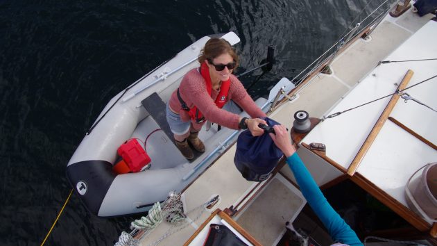 A woman wearing sunglasses standing in a dinghy passing up a dry bag to someone on the deck of a yacht