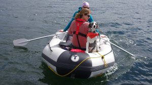 two women rowing a dinghy with a dog in a lifejacket